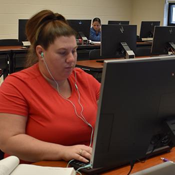 Female Student in computer lab wearing headphones with student on computer in background